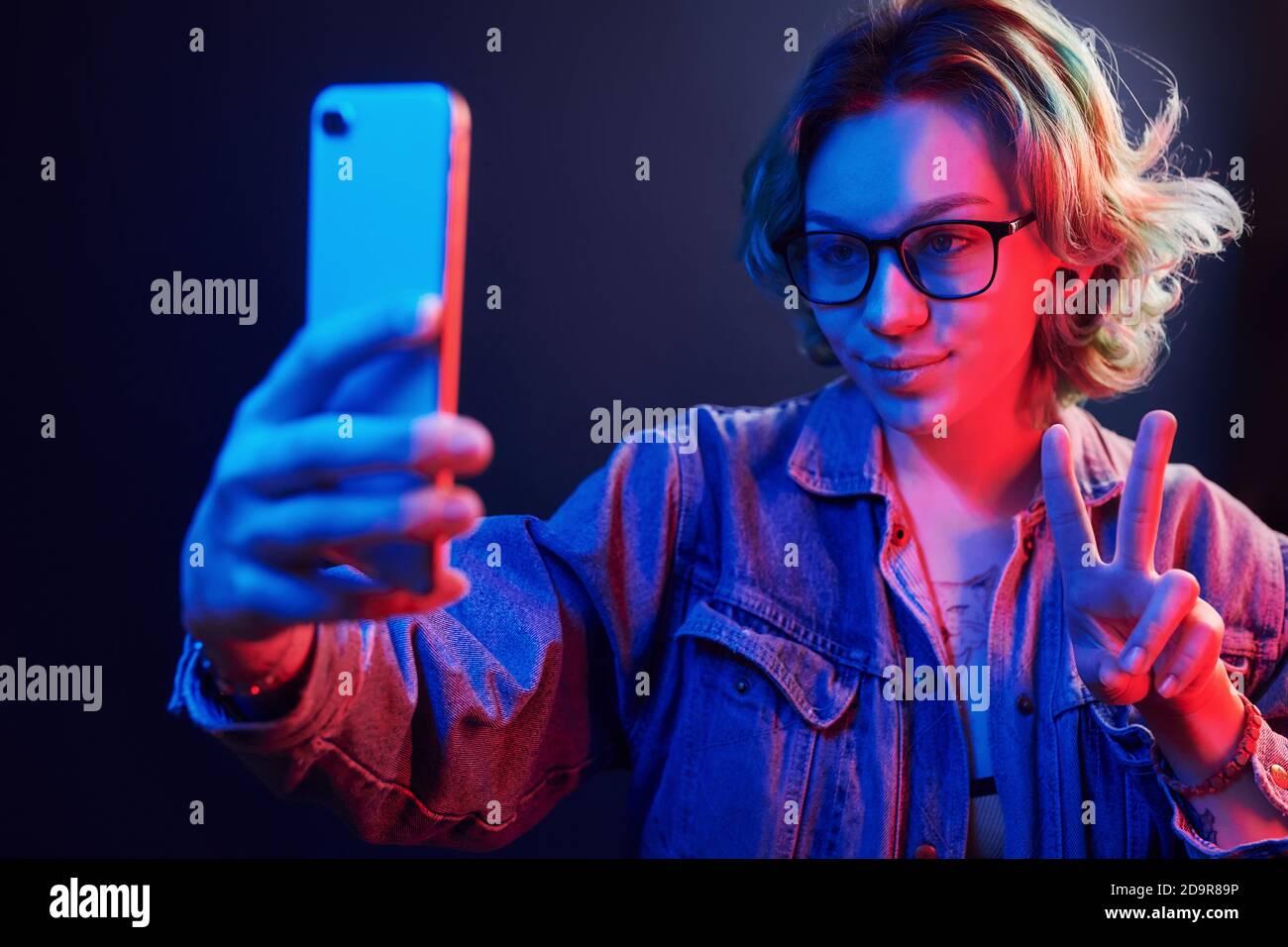 Ritratto di giovane ragazza alternativa in occhiali con capelli verdi che fare selfie per telefono in rosso e blu neon luce in studio Foto Stock
