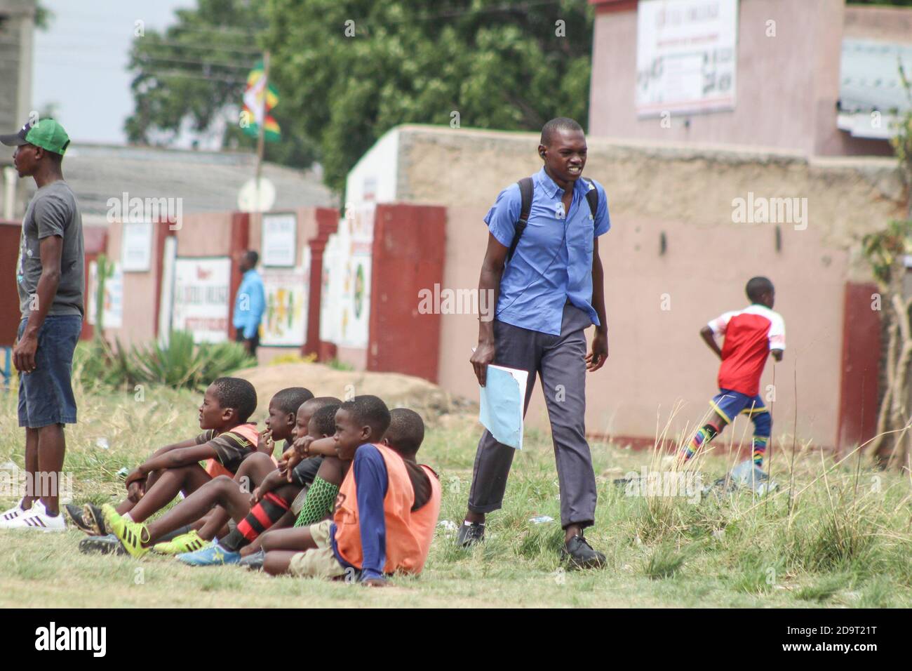 Passa un allievo in uniforme mentre i giocatori della junior accademia celebrano un gol durante un torneo di calcio u12 a Chitungwiza, Zimbabwe. Il paese si prepara per l'apertura del resto dei voti a scuola. I bambini sono stati a casa da marzo, quando è stato annunciato il blocco a causa della covid 19. Zimbabwe. Foto Stock