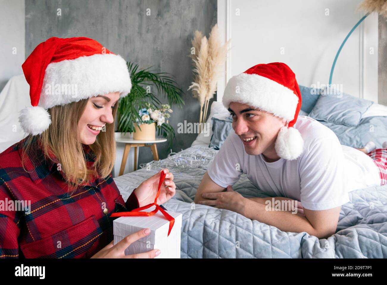 Natale, vacanze, inverno e concetto di gente. Felice coppia con confezione regalo a casa indossando cappelli santa. Giovane uomo sorridente sdraiato sul letto dare Foto Stock