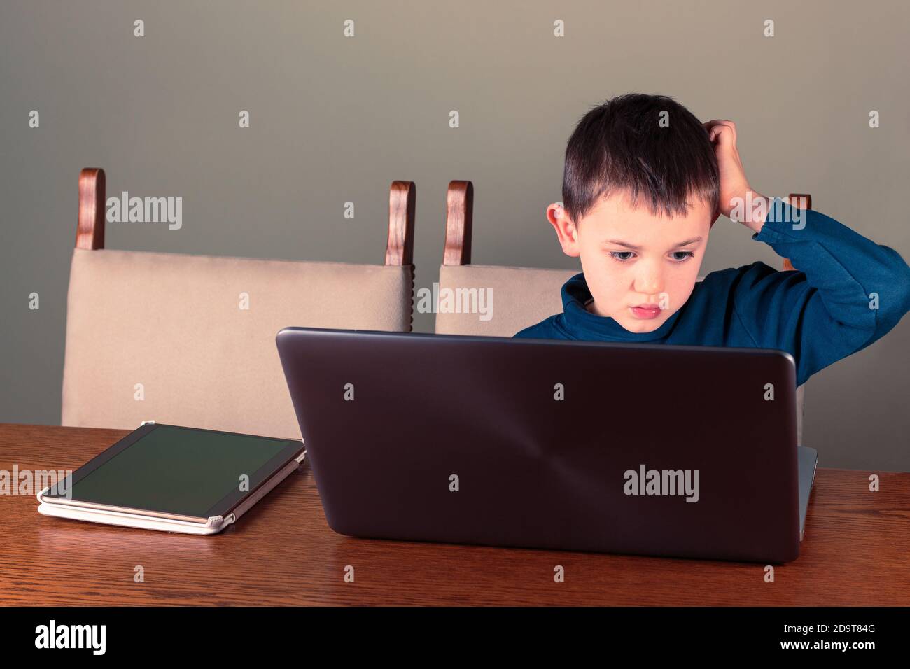 Il ragazzo di otto anni che usa computer portatili e tablet gli graffia la testa. Homeschooling o tema della tecnologia digitale Foto Stock