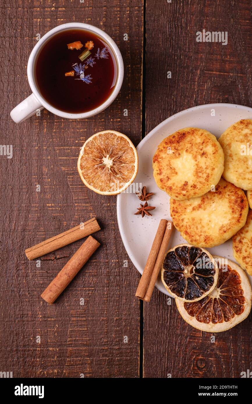 Frittelle di formaggio cottage con tè nero aromatico caldo, umore di prima colazione di Natale con anice, cannella e agrumi secchi su sfondo di legno, vista dall'alto Foto Stock