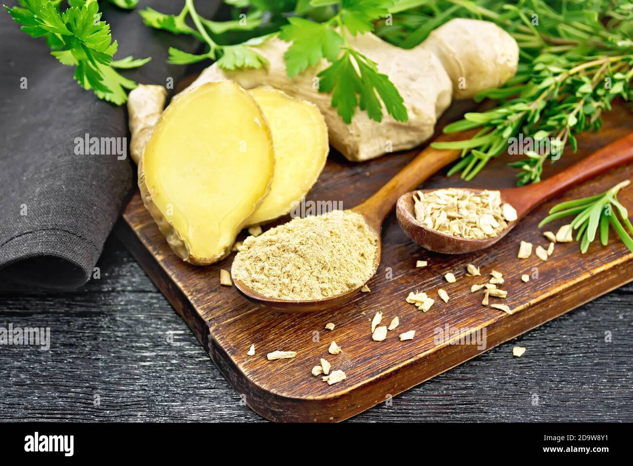 Zenzero macinato e fiocchi in due cucchiai di legno, radice di zenzero, erbe speziate, tovagliolo su uno sfondo di legno nero Foto Stock