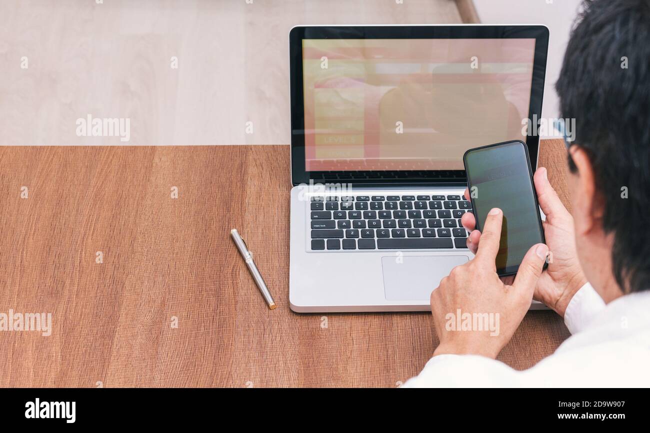 Prendere da un uomo con la sua schiena girata che sta navigando in internet sul suo cellulare e laptop su una scrittura in legno nel suo ufficio. Applicazioni che un Foto Stock