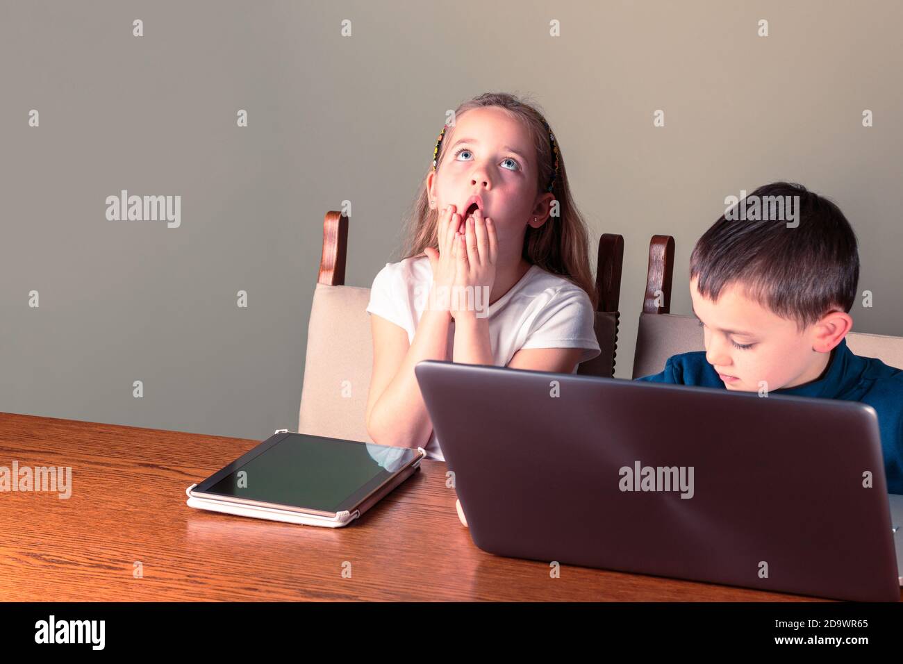 Carino bambini che usano il computer portatile e tablet, la ragazza guarda disperatamente in su tenendo le mani sulla bocca; homeschooling o tema di tecnologia Foto Stock