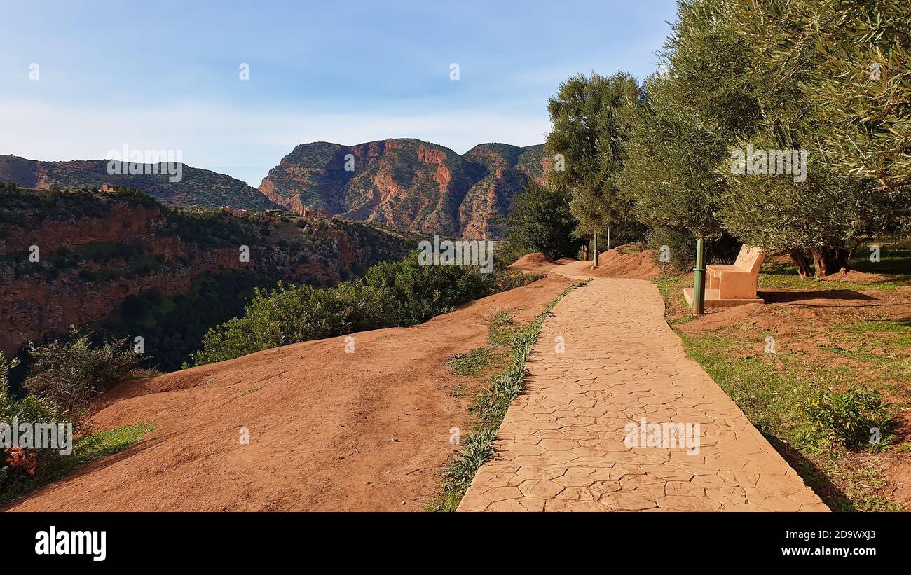 Sentiero ai margini di un canyon sopra le cascate Ouzoud a Ouzoud, Marocco, con una panca di riposo e le montagne sullo sfondo in giornata di sole. Foto Stock
