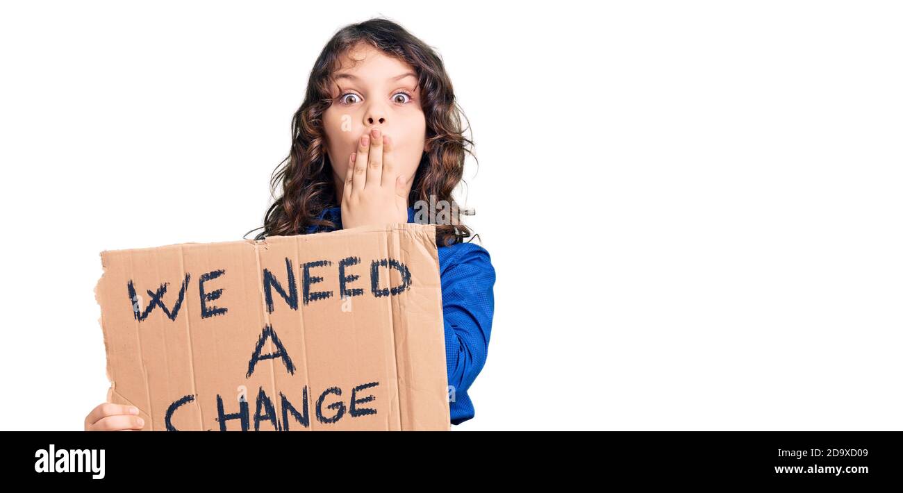 Cute bambino ispanico con i capelli lunghi che tengono abbiamo bisogno di un cambio banner che copre la bocca con la mano, scioccato e impaurito per errore. Sorpresa espressione Foto Stock