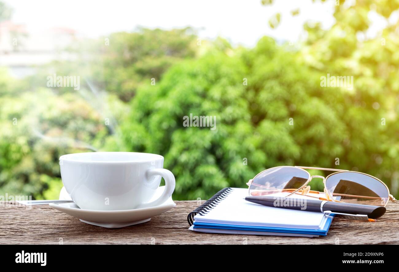 Tazza di caffè calda con un notebook, occhiali da sole e penna sul vecchio pavimento in legno con colorato sfondo verde natura. Foto Stock