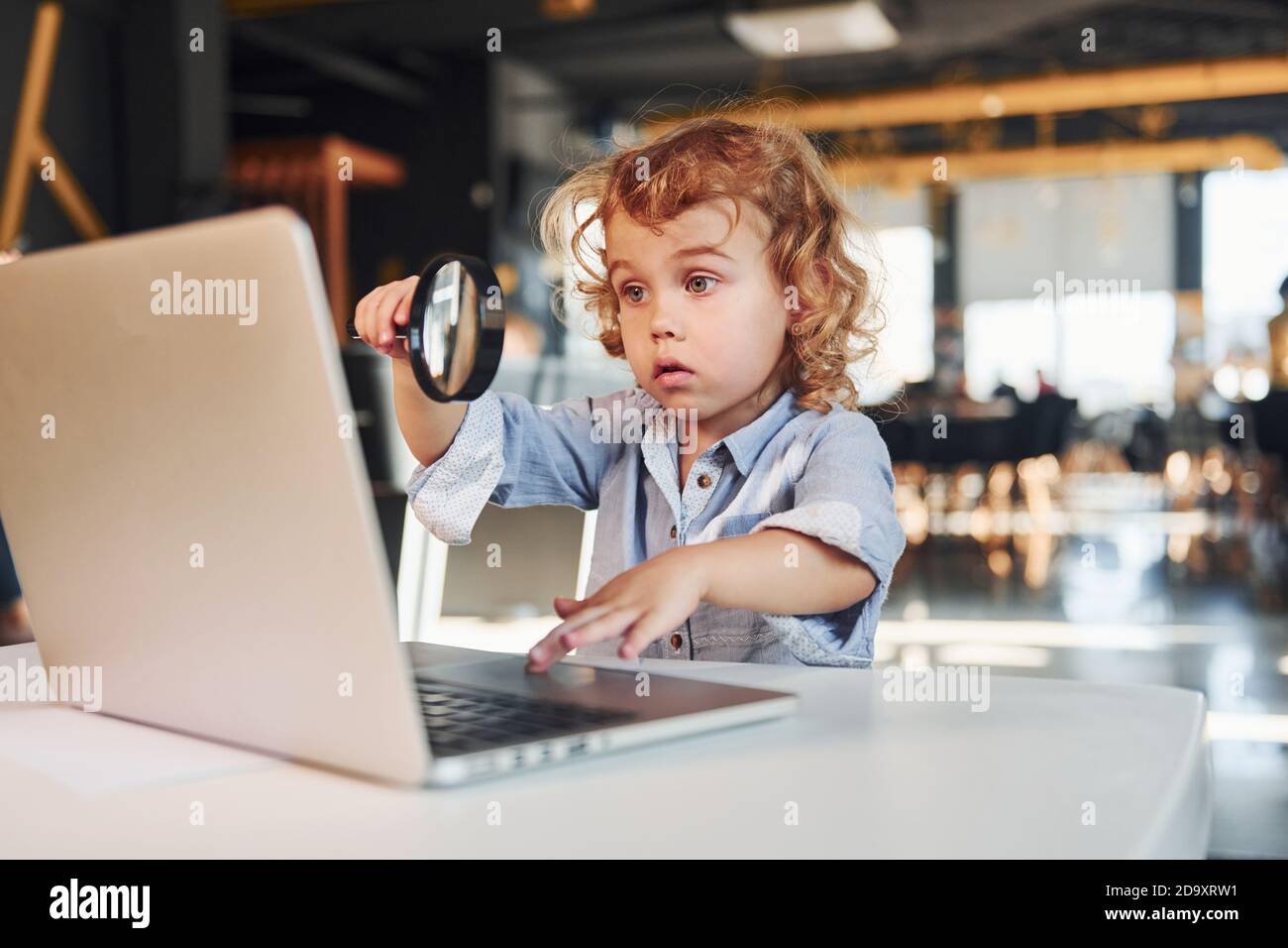 Bambino intelligente in abiti casual con computer portatile sul tavolo hanno divertimento con la lente d'ingrandimento Foto Stock