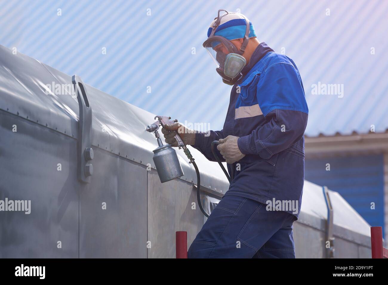 Lavoro industriale. Adescamento di prodotti metallici dalla pistola di compressione. Un operatore in tute e un respiratore dipinge il corpo di un rimorchio o di un Foto Stock