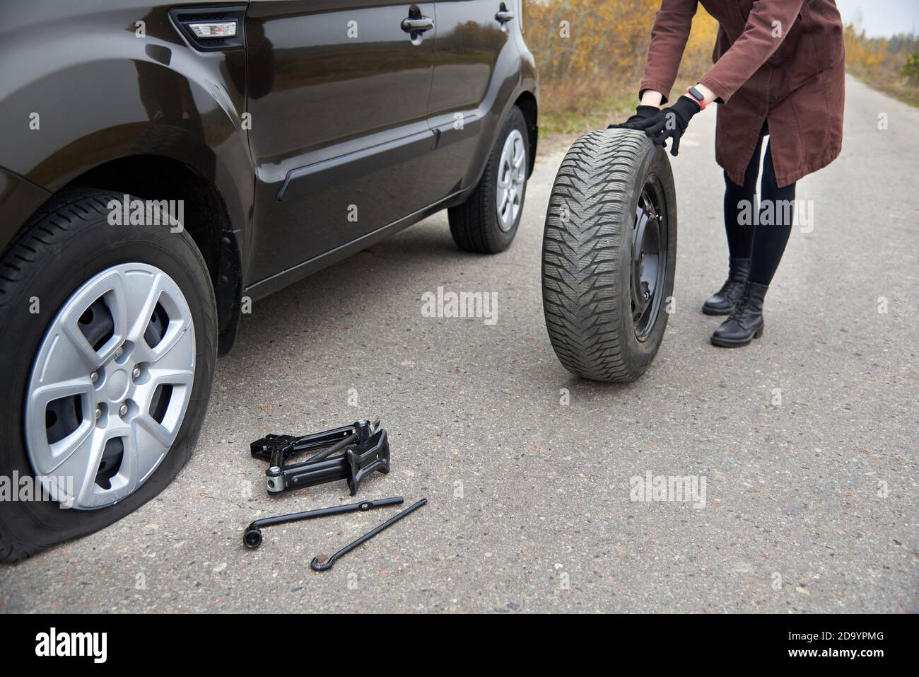 Una giovane donna rotola il pneumatico di scorta vicino alla sua auto con uno pneumatico sgonfio, difficoltà sulla strada. Foto Stock