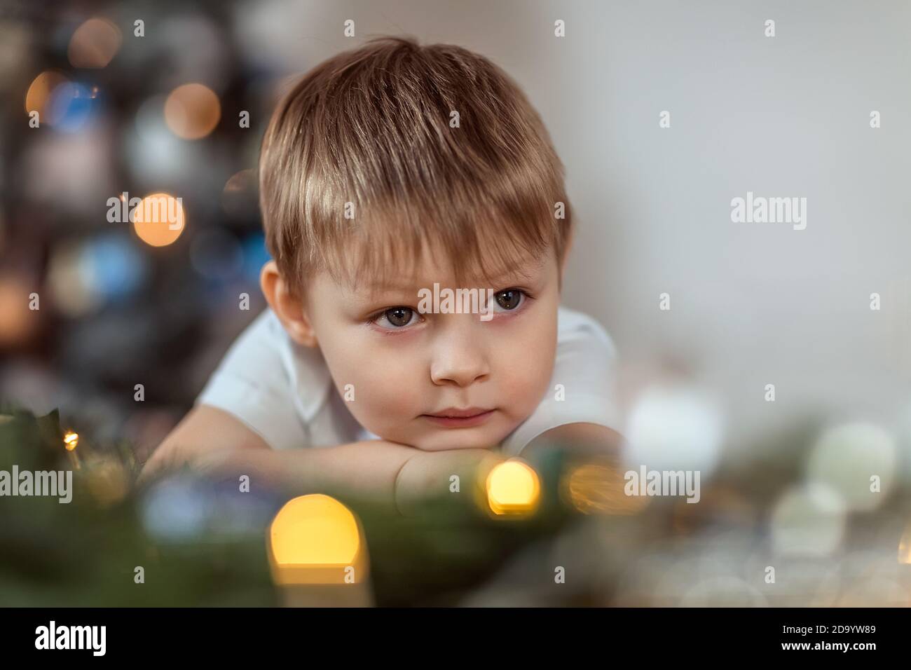 Un bel ragazzo di 4-5 anni è seduto o in piedi vicino all'albero di Natale. Sogna una vacanza e dei regali, fa un desiderio. Il concetto di wi Foto Stock