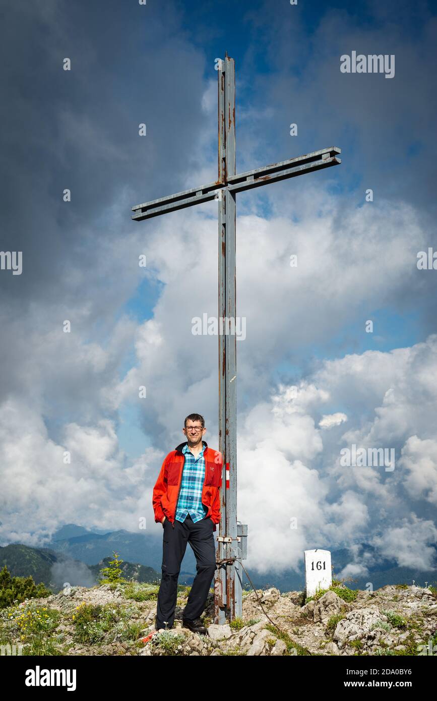 L'escursionista maschile si trova accanto alla croce sulla cima del Monte Halserspitze nelle Alpi Bavaresi, in Germania Foto Stock