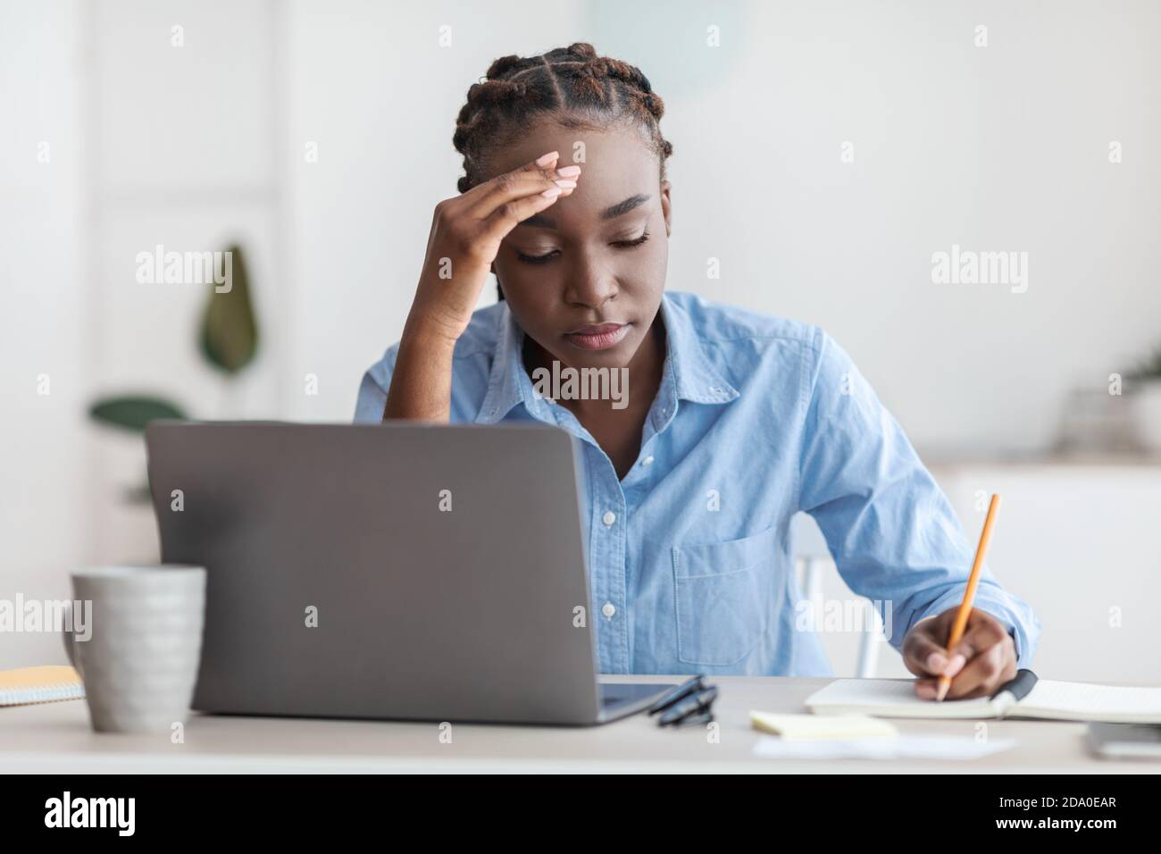 Stanco dipendente nero dell'ufficio seduto alla scrivania con il computer portatile, prendendo appunti Foto Stock