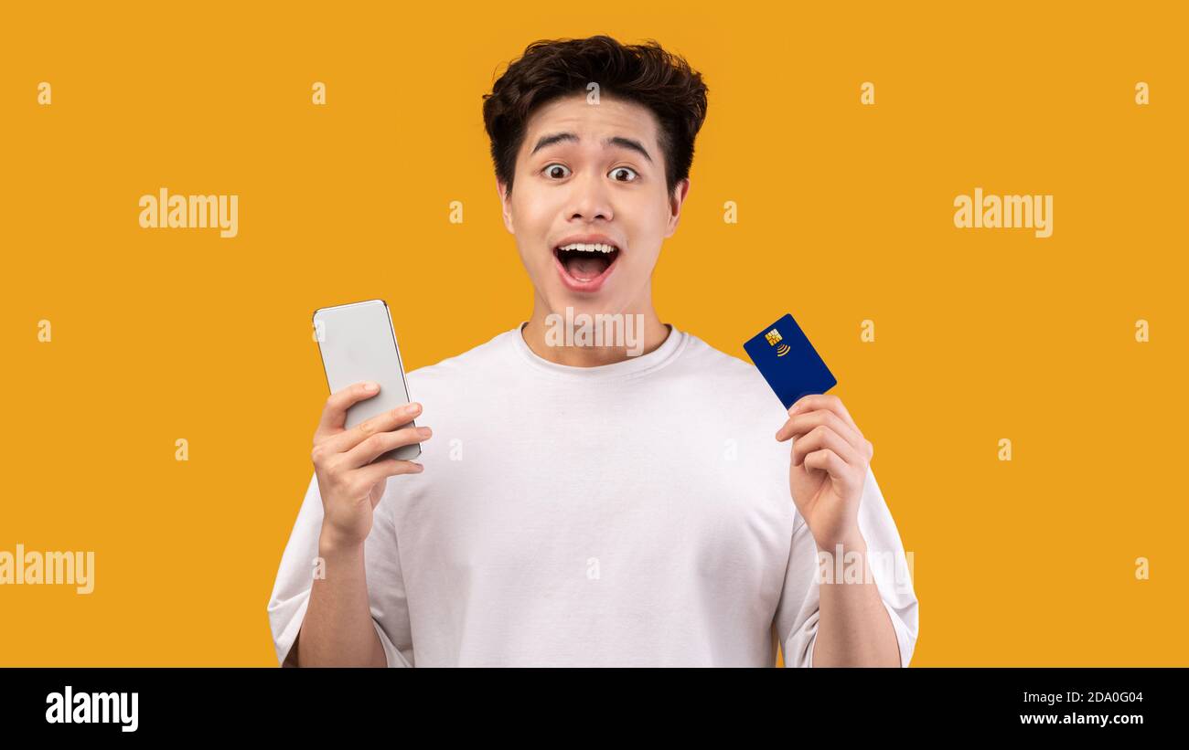 Ragazzo asiatico eccitato che tiene la carta di credito e il cellulare in studio Foto Stock