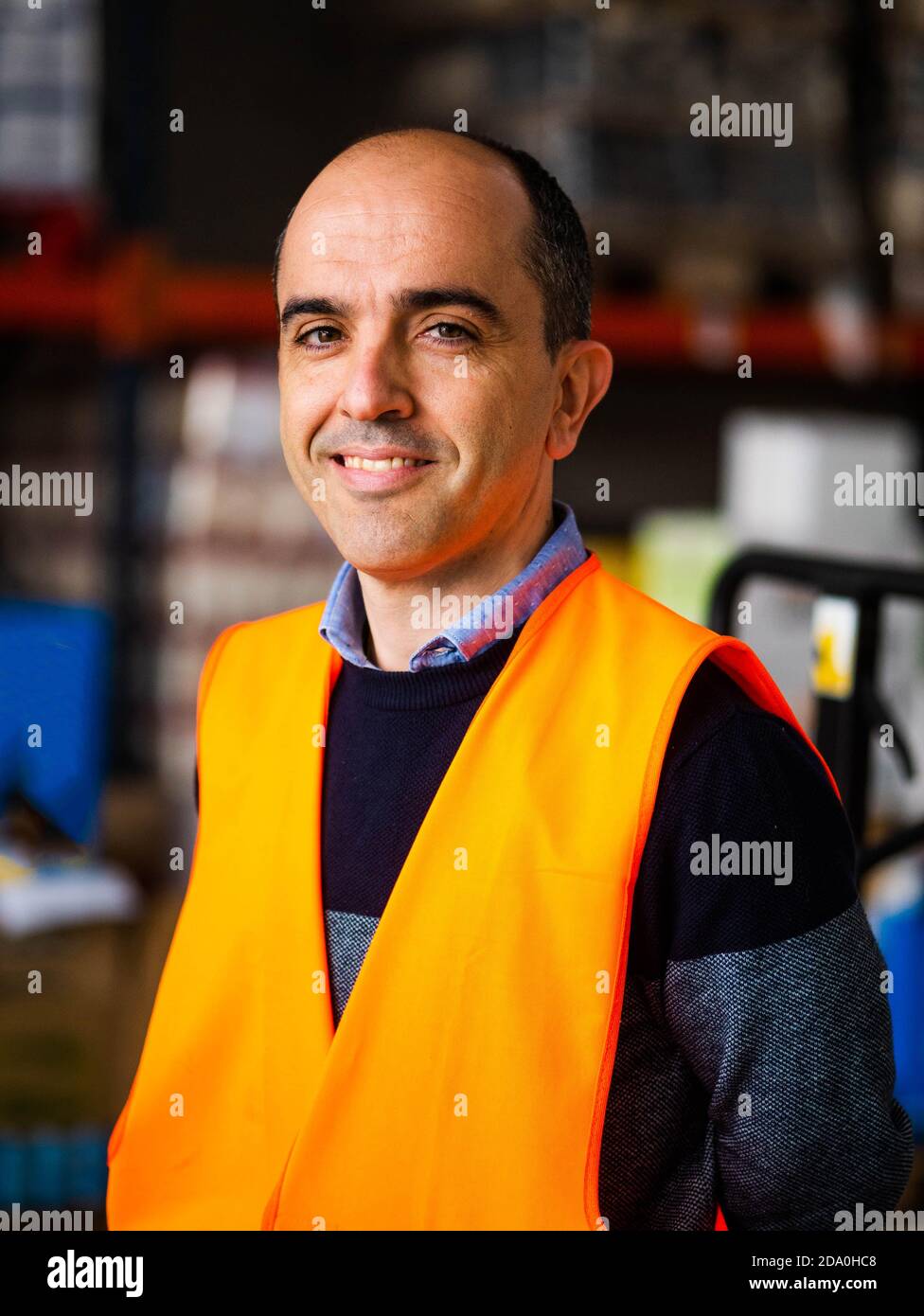 Felice adulto lavoratore maschio che indossa giubbotto arancione ad alta visibilità in piedi vicino ai rack del magazzino e guardando la telecamera Foto Stock