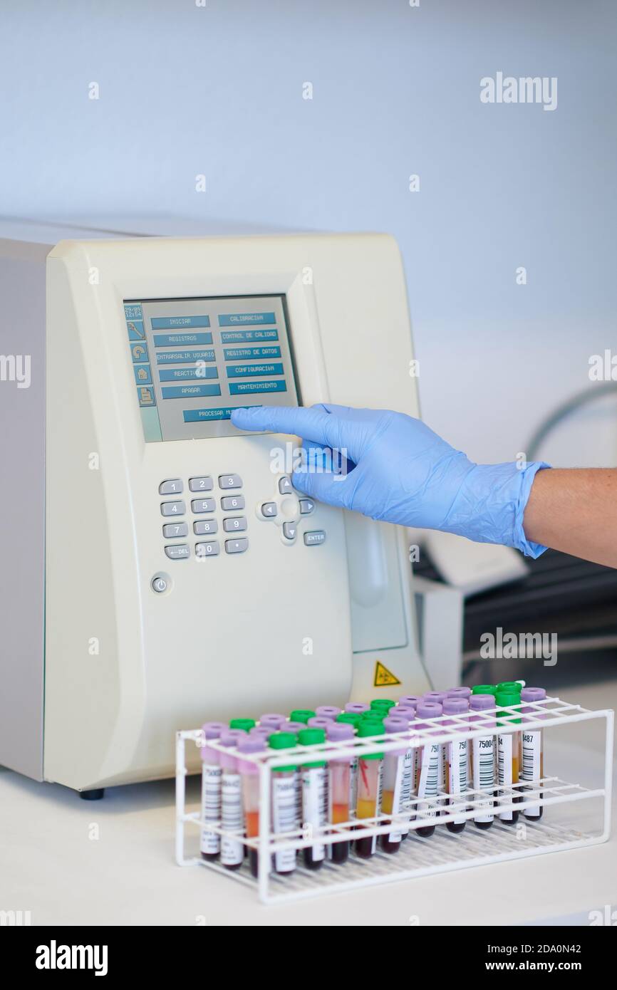 Coltivare un tecnico medico irriconoscibile che tocca il display dell'analizzatore ematologico vicino tabella con provette per sangue in laboratorio Foto Stock