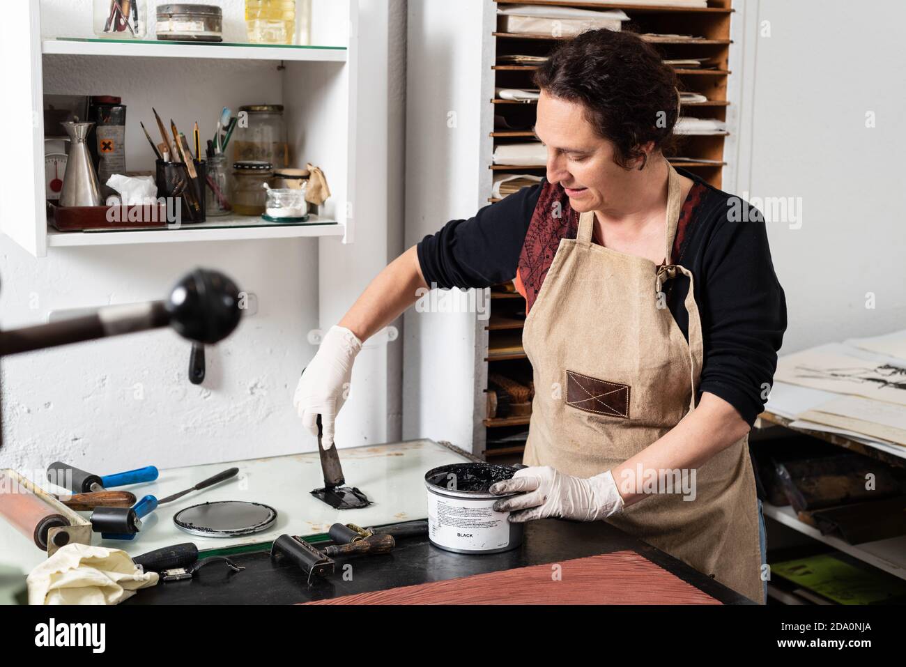 Master professionale femminile in grembiule e guanti con inchiostro nero per la creazione di rilievi di stampa durante il lavoro in officina di incisione Foto Stock