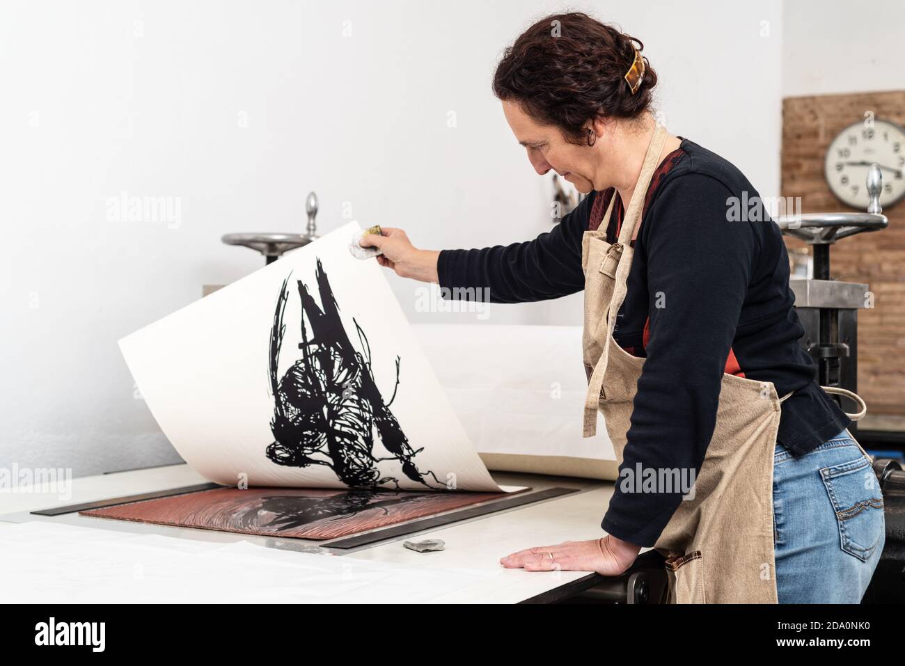 Vista laterale di artista femminile esperto che rimuove il foglio di carta con impronta dal pezzo di linoleum collocato sulla macchina da stampa mentre lavorare in creatività Foto Stock