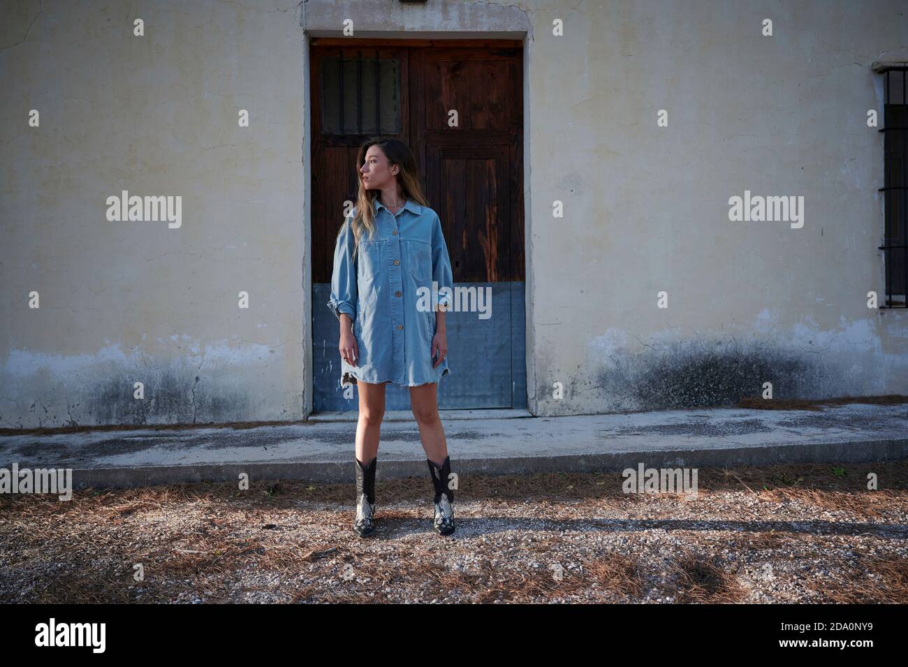 Corpo pieno di giovane donna slim in elegante abito in denim e stivali in piedi su prato erboso coperto di brina contro stagionato casa rurale in autunno Foto Stock