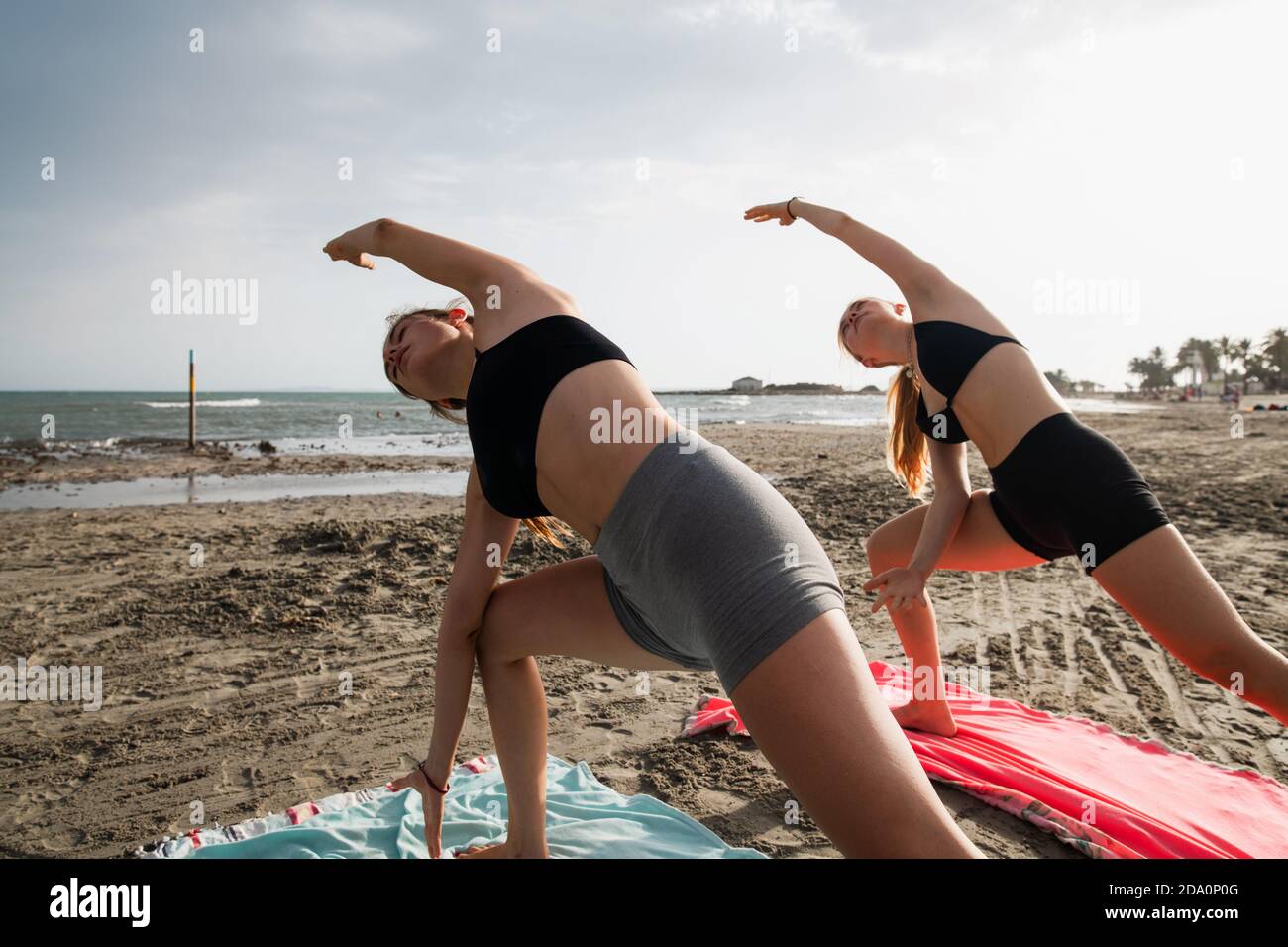 Vista laterale di donne gravi e flessibili che praticano yoga su sabbia costeggia vicino al fiume che si increspa in un giorno cupo Foto Stock