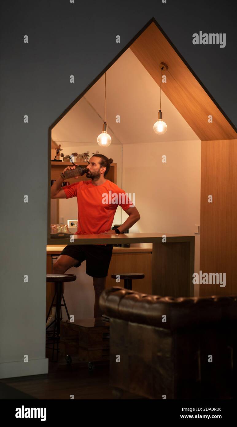 Sportivo sicuro con la mano sulla vita in piedi in cucina e. acqua potabile dopo l'allenamento in palestra Foto Stock