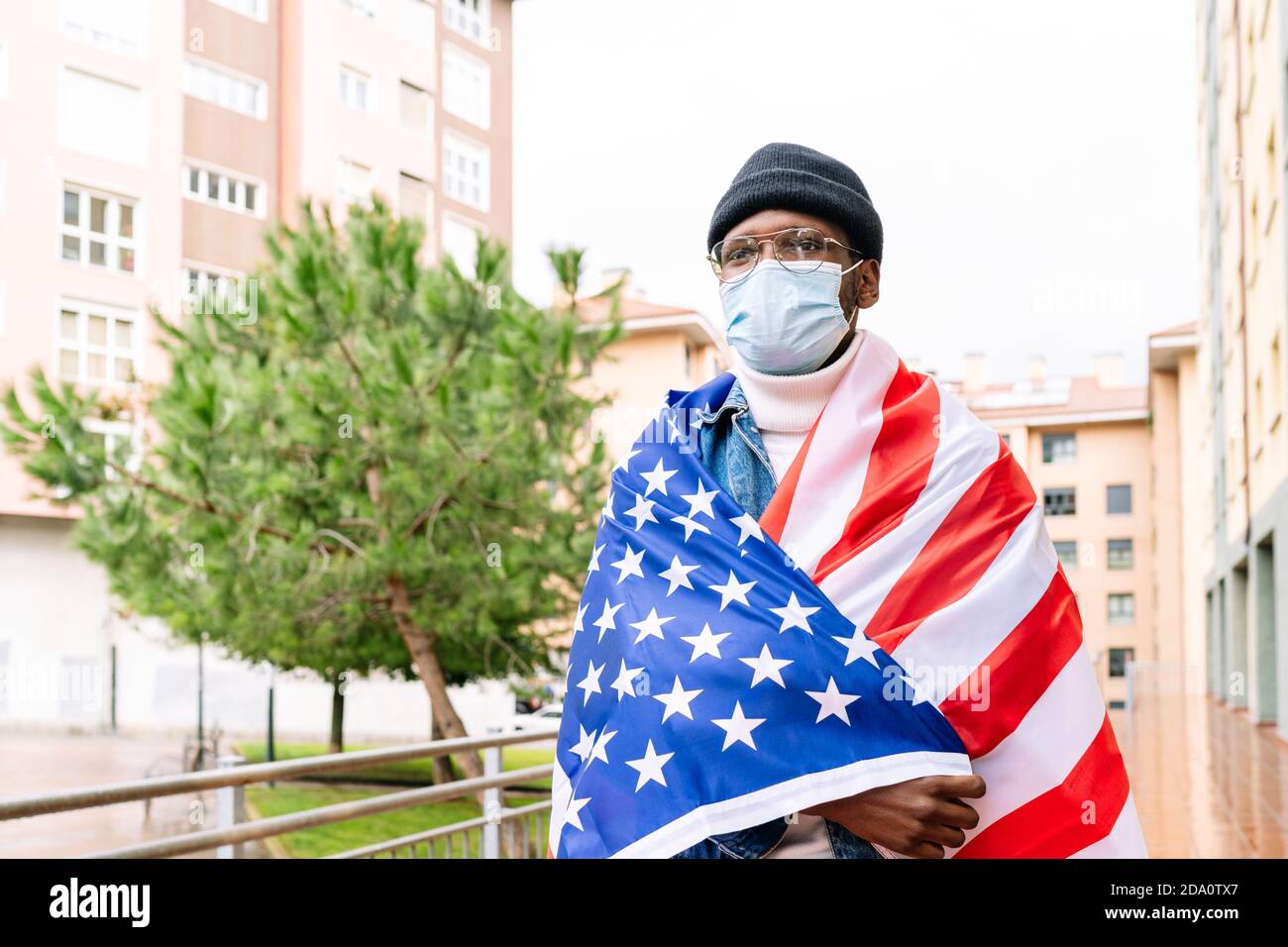 Uomo patriottico afroamericano in maschera medica avvolta negli Stati Uniti bandiera nazionale in piedi sulla strada durante la pandemia di coronavirus e guardare via Foto Stock