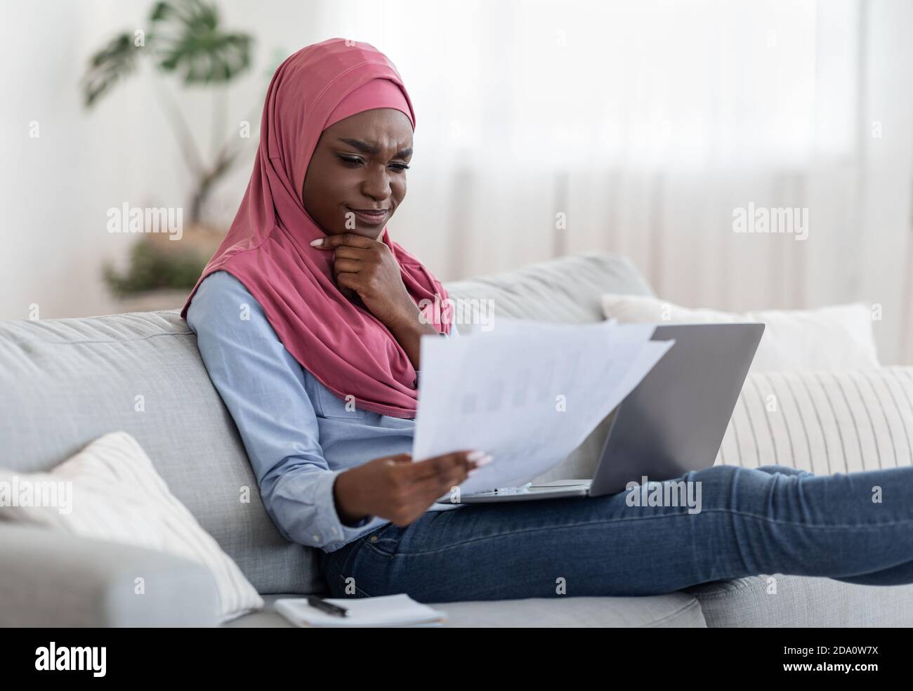 Donna musulmana nera premurosa che controlla i documenti e lavora sul computer portatile a casa Foto Stock