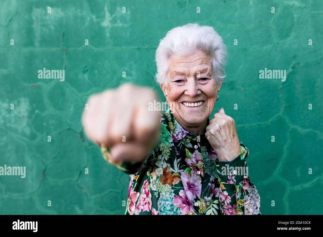 Allegro anziano donna capelli grigi in blusa colorata ed elegante che ha divertimento e pugilato alla macchina fotografica mentre si sta contro il muro verde Foto Stock