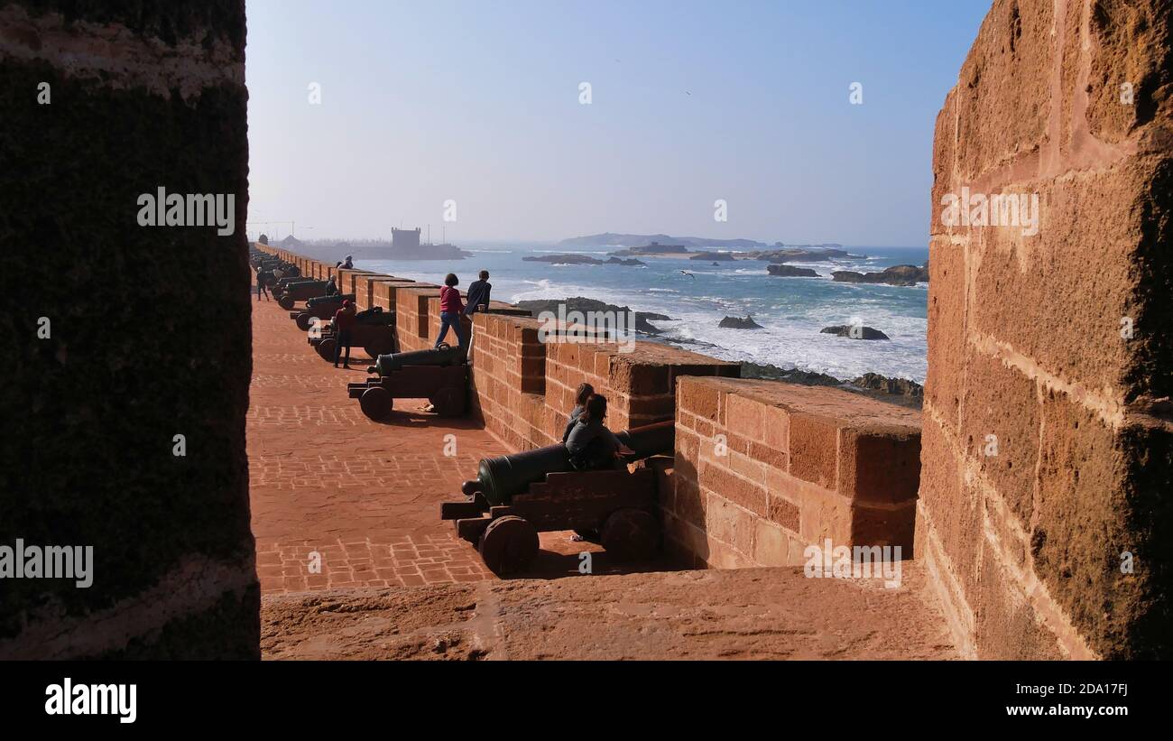I turisti seduti su vecchi cannoni e godendo la brezza marina salata del selvaggio oceano Atlantico sulla parete storica nella Medina di Essaouira, Marocco. Foto Stock