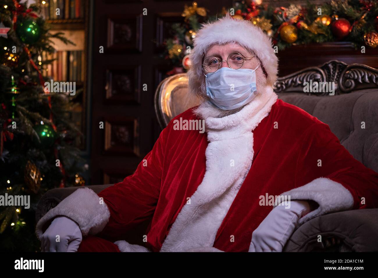 Babbo Natale in maschera medica. Ritratto positivo Santa seduta vicino albero di Natale. Felice anno nuovo Covid 2021 Foto Stock