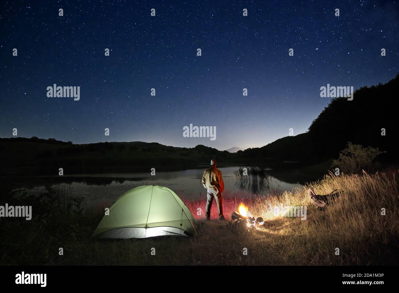 Uomo in piedi vicino a tenda illuminata e falò vicino al lago sotto cielo stellato nel Parco dei Nebrodi, Sicilia Foto Stock
