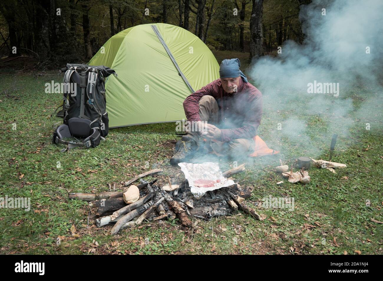 Uomo seduto sull'erba del Parco dei Nebrodi e cucina carne e funghi sul fuoco vicino tenda e zaino, Sicilia Foto Stock
