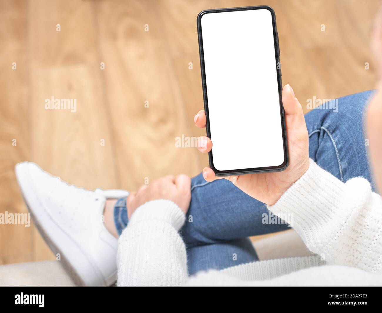 mockup cellulare. donna tiene un cellulare mockup. La donna si siede a croce in un pullover bianco e jeans su un divano e mostra il telefono cellulare con una w vuota Foto Stock