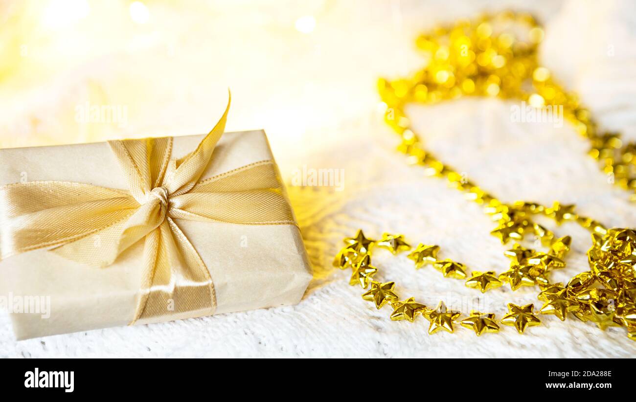 Scatola con regalo di Natale in carta chiara è legata con un nastro d'oro su sfondo bianco lavorato a maglia con decorazione. Golden ghirland di stelle. Anno nuovo. Congratul Foto Stock