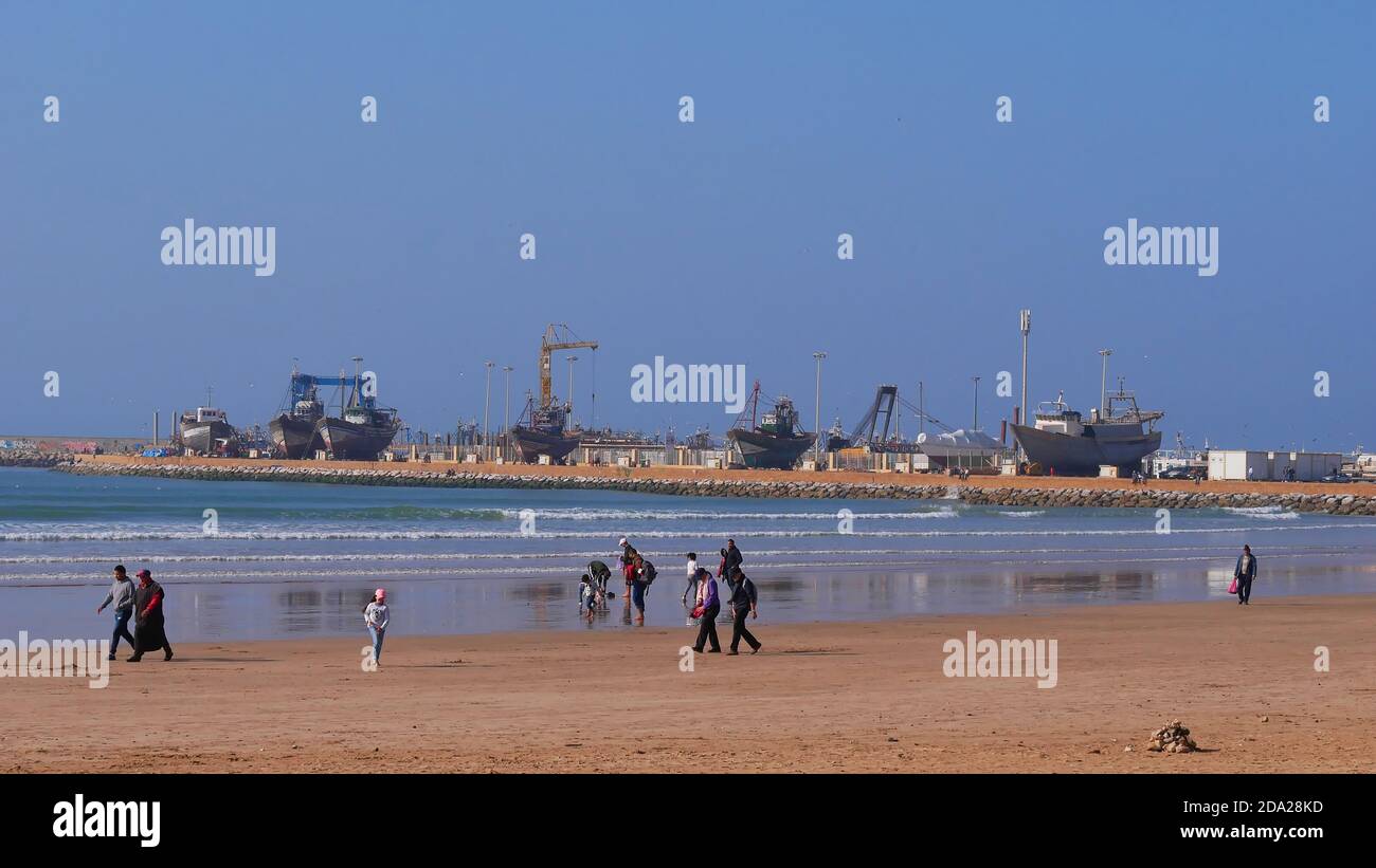 Essaouira, Marocco - 01/04/2020: Persone che si godono la passeggiata sulla famosa spiaggia di Tagharte al ventoso Oceano Atlantico con le barche da pesca jacked. Foto Stock