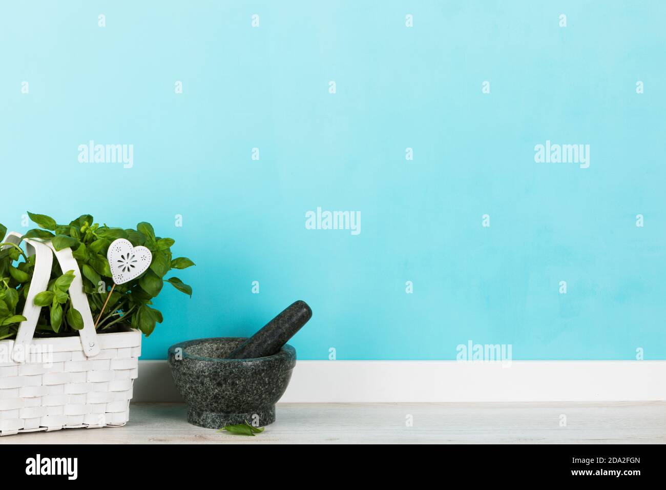 Fondo cucina con pianta di basilico, in cesto bianco, mortaio e pestello di fronte al muro dipinto turchese Foto Stock