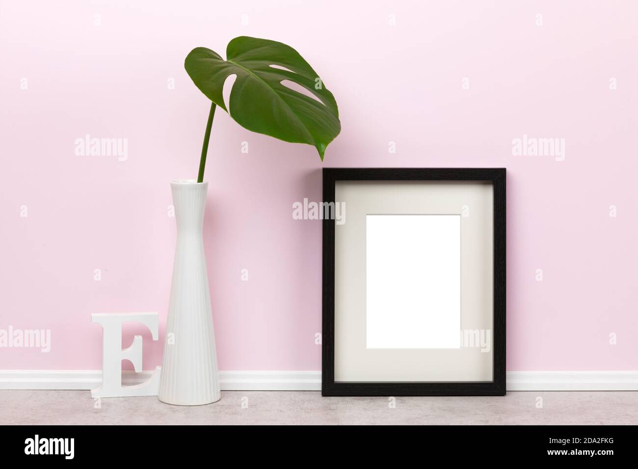 Elegante dima mockup con cornice verticale in legno nero, con lettera e in legno opaco e foglia di monstera deliziosa in vaso davanti alla parete rosa Foto Stock