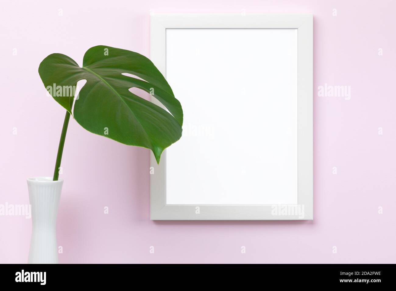 Mascherina verticale bianca del mockup del quadro sulla parete rosa con vaso e foglia di monstera davanti, area vuota del quadro isolata con tracciato di ritaglio Foto Stock