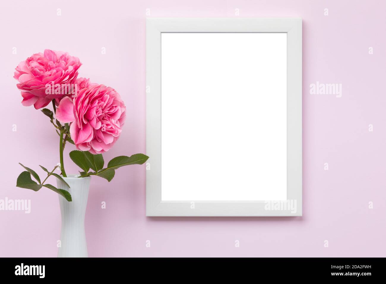 Mascherina verticale bianca del mockup della cornice dell'immagine sulla parete rosa con vaso e rose davanti, area vuota dell'immagine isolata con il tracciato di ritaglio Foto Stock