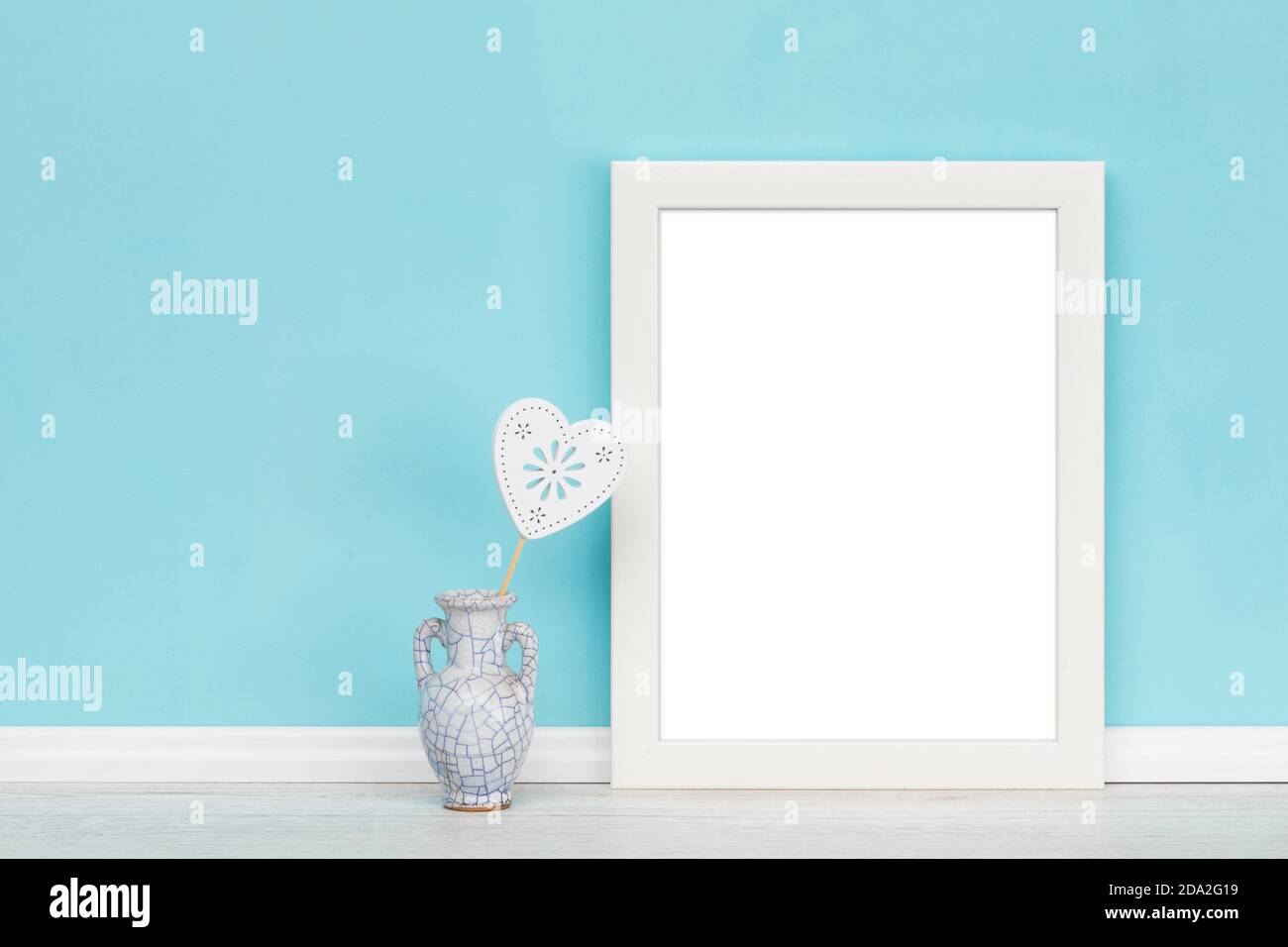 Modello mockup di sfondo con cornice verticale in legno bianco vuoto e vaso con decorazione a forma di cuore di fronte alla parete turchese, grammo interno Foto Stock