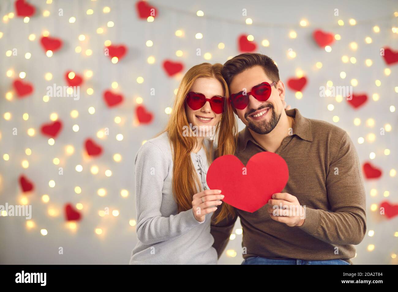 Giovane felice coppia amorevole in occhiali a forma di cuore in piedi e. tenendo il cuore rosso grande nelle mani Foto Stock