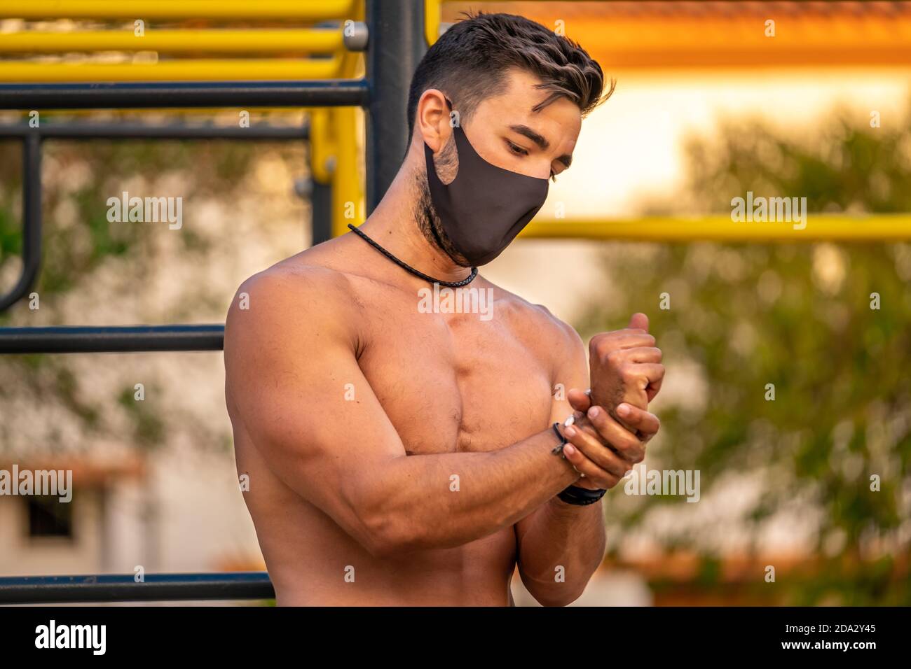 Giovane latino senza camicia e pantaloni neri e. maschera disinfettando le mani con gel idroalcolico durante l'allenamento un parco calistenico Foto Stock