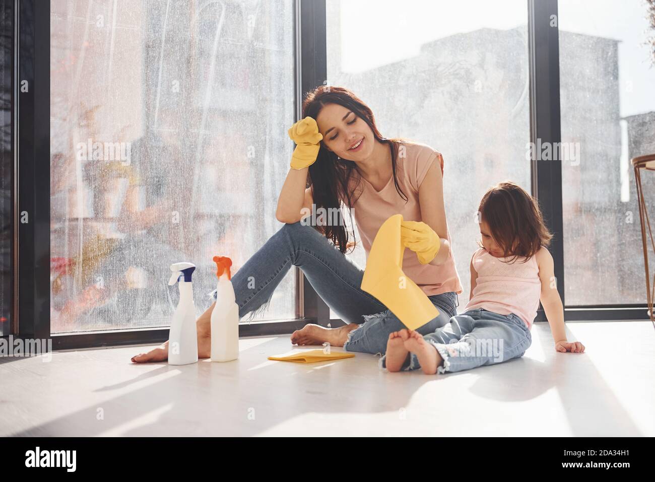 Donna stanca con sua figlia si siede sul davanzale della finestra con bottiglie di spray detergente e con guanti Foto Stock