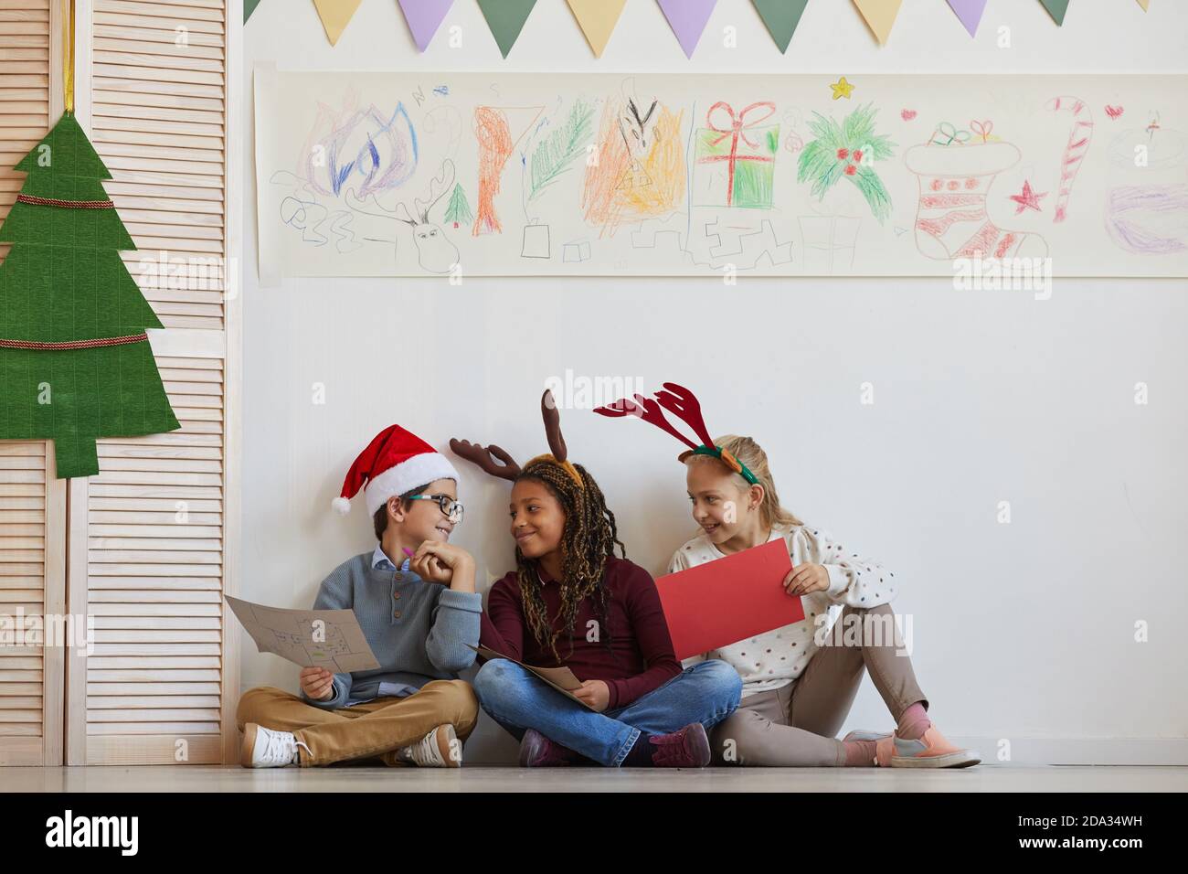 Ritratto a lunghezza intera di un gruppo multietnico di bambini seduti sul pavimento che tengono le foto mentre si gode la lezione d'arte di Natale, copy space Foto Stock