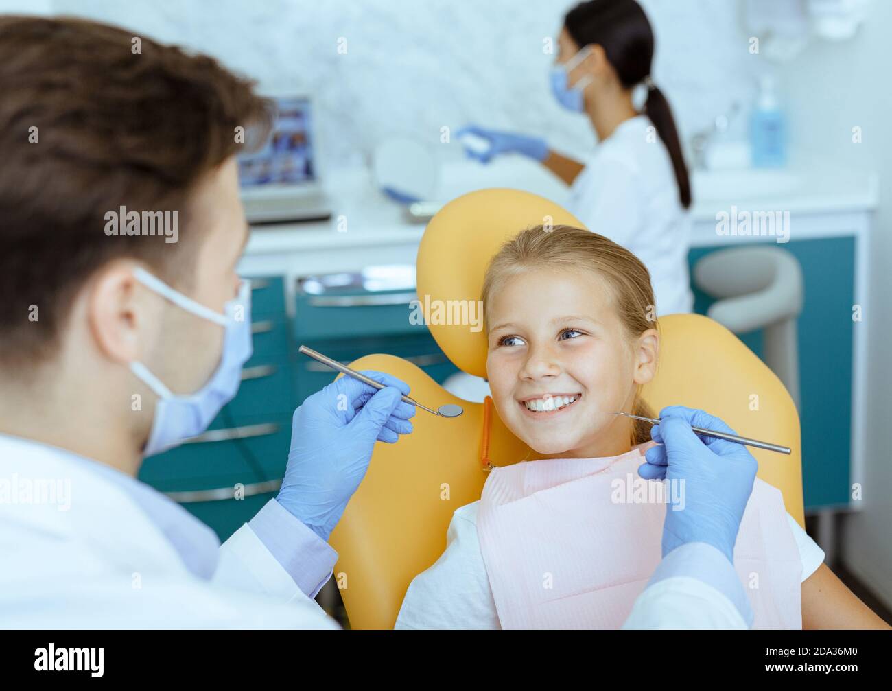 Medico maschile in camice bianco con guanti di gomma e protettivo la maschera tratta i denti della ragazza sorridente in sedia Foto Stock