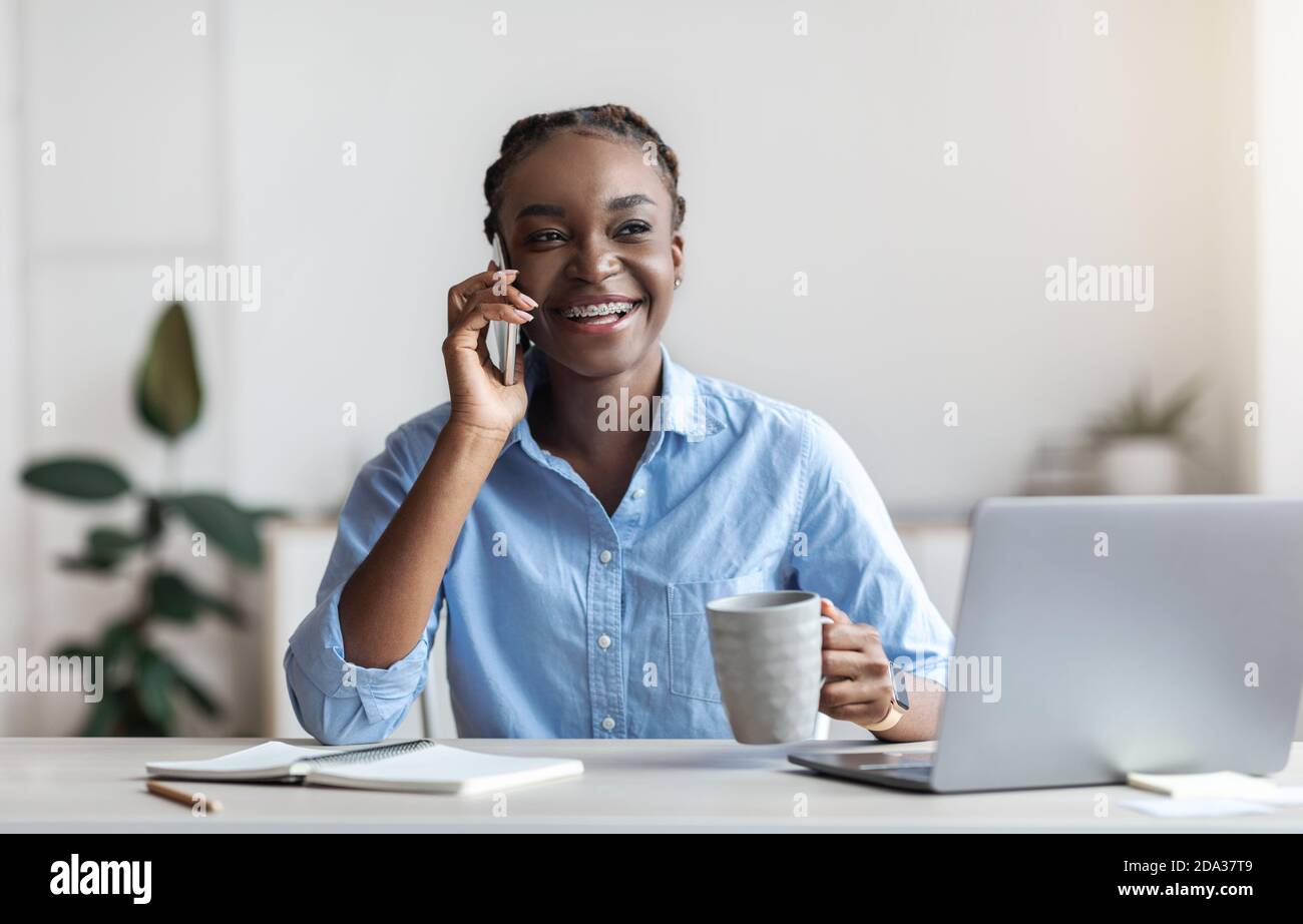 Young Black Female Office Employee Relaxing at Workplace with Cellphone E caffè Foto Stock