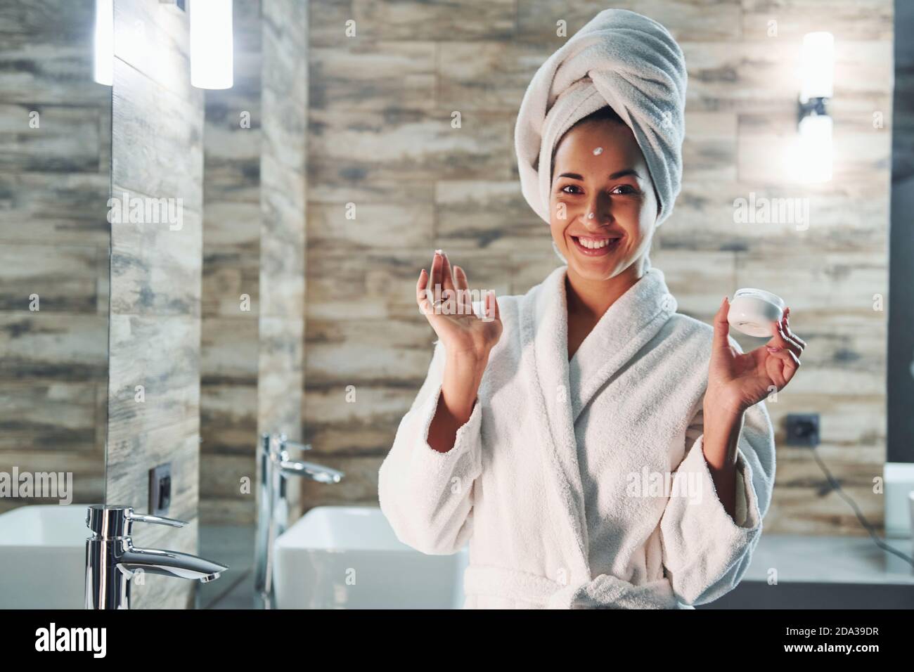 Bella giovane donna in asciugamano bianco in piedi in bagno e. prendendosi cura del suo viso Foto Stock