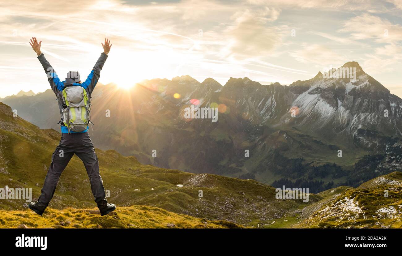 Uomo Hiker di successo con zaino che salta con le braccia sollevate. Incredibile alba in montagna con belle lenti e raggi solari. Alpi, Allgau Foto Stock