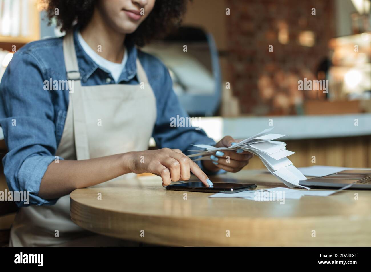 Occupato african american giovane proprietario di signora in grembiule lavora con le carte, conta i costi e i profitti in interno Foto Stock
