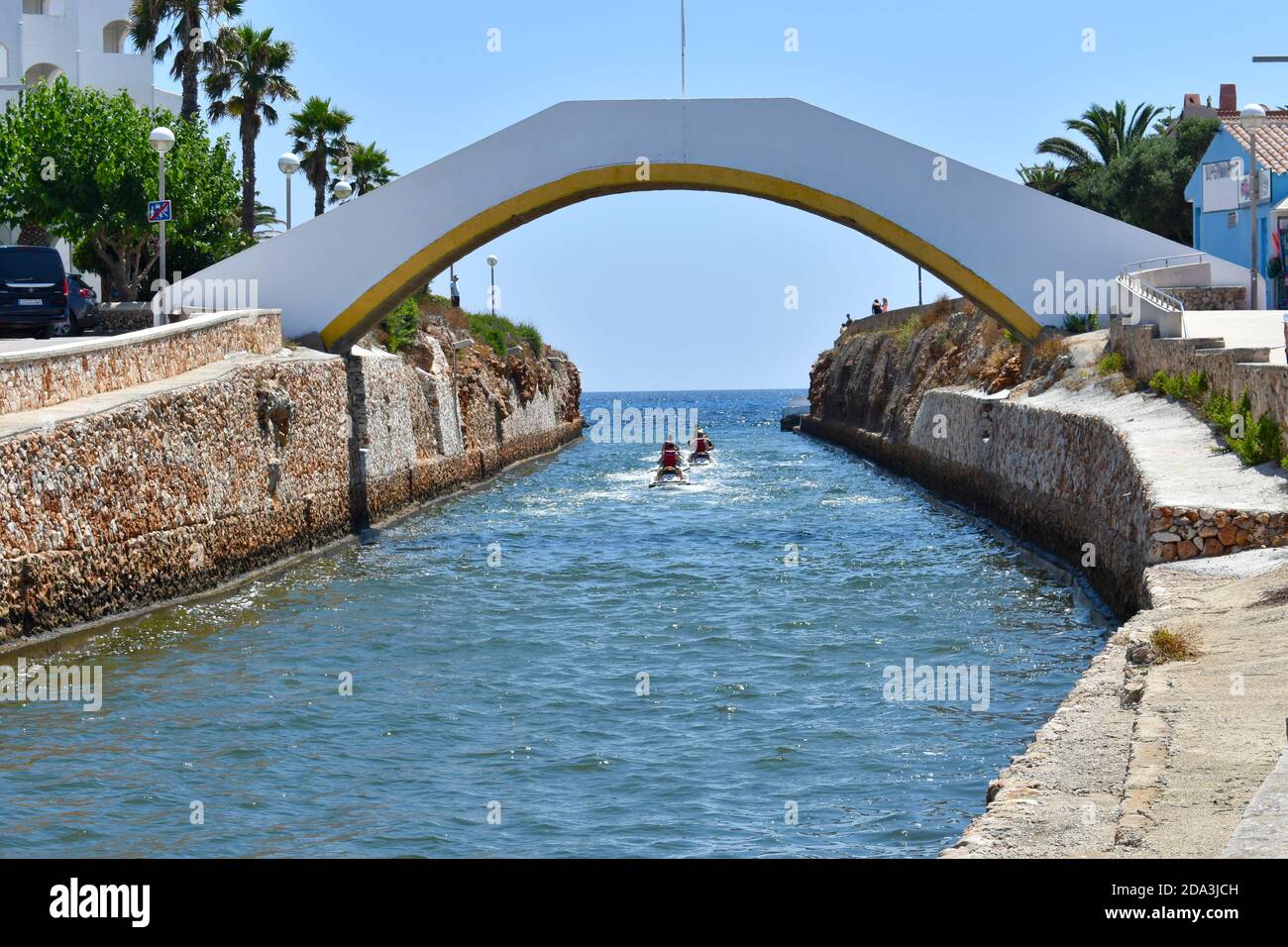 ponte sul punto di sbocco al mare a Minorca con barche da sci d'acqua che escono al mare sotto il ponte. Pareti adiacenti fiancheggiate da palme Foto Stock